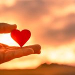 Hand holding wooden heart at beautiful sunset.